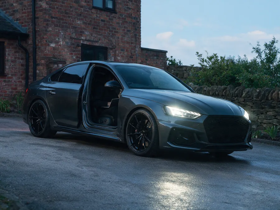 Grey BMW M3 parked outside a brick house at dusk with the driver door open and the headlights on